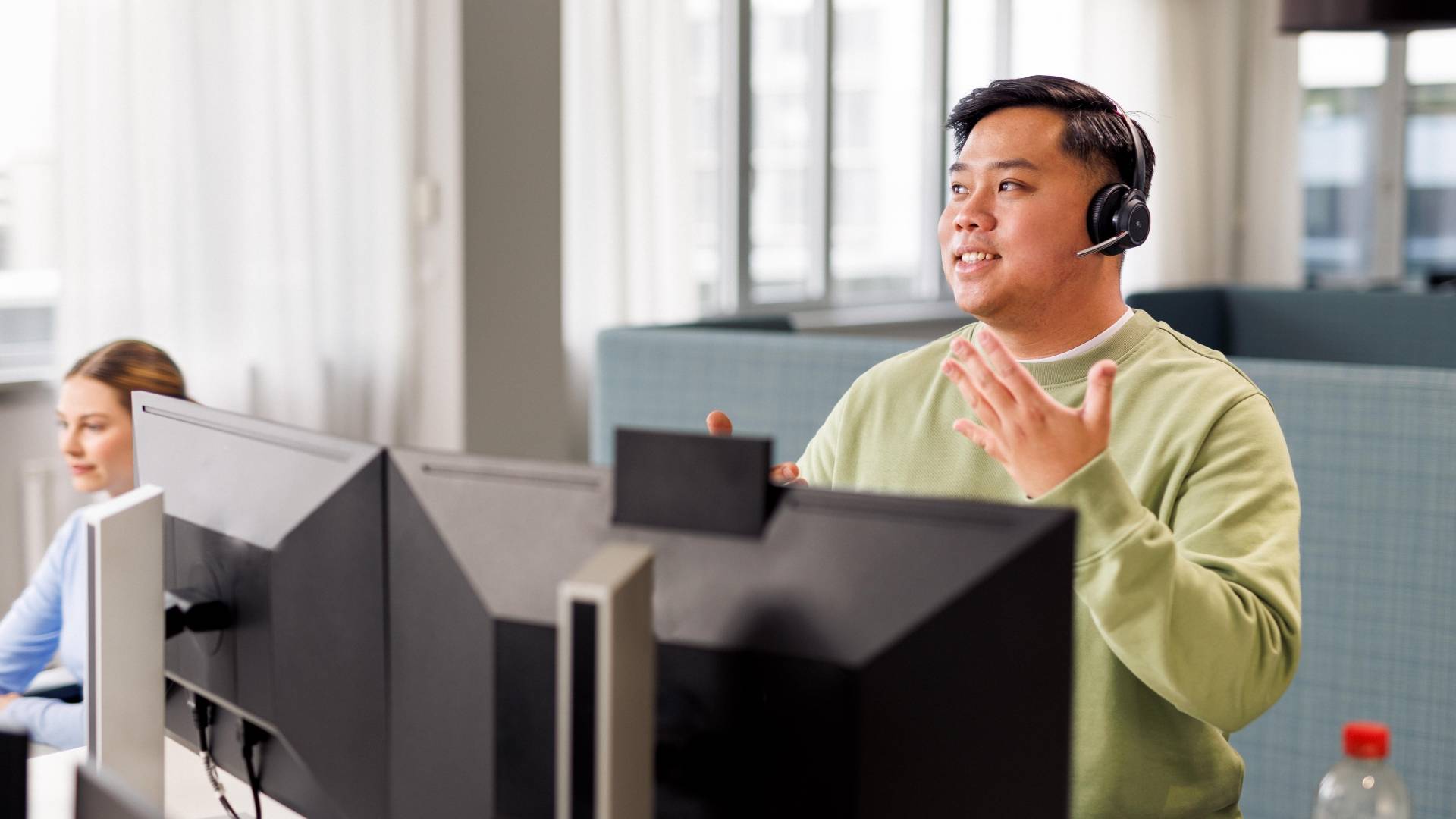 Ein Azubi sitzt mit Headset und im hellgrünen Pullover vor einem Computer. Er führt ein Gespräch.