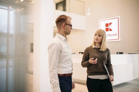 Eine Mitarbeiterin ist am Empfang mit ihrem Kollegen im Gespräch. Sie stehen vor einem weißen, modernen Tresen. Im Hintergrund ein großes Kaufland-Logo.