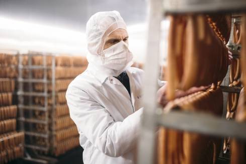 Ein Teamleiter mit Mundschutz und in Schutzkleidung vor einem Regalstand mit aufgehangenen Würsten bei der Qualitätssicherung im Kaufland-Fleischwerk.