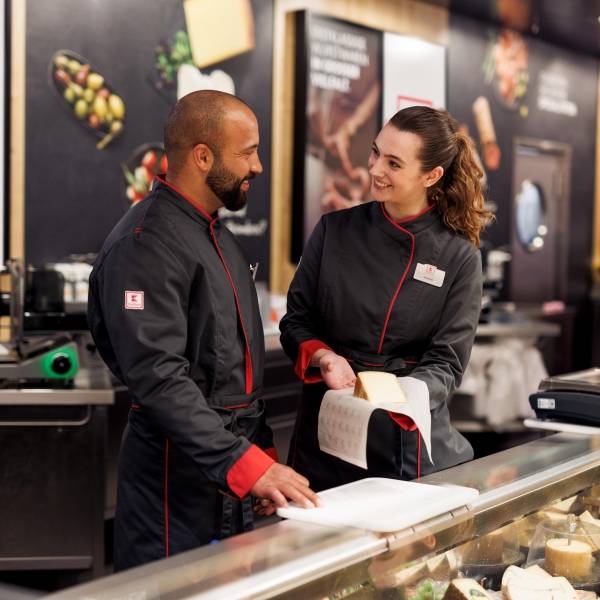 Eine Mitarbeiterin und ein Azubi in schwarzem Kaufland-Overall stehen hinter der Käsetheke. Die Mitarbeiterin erklärt etwas.