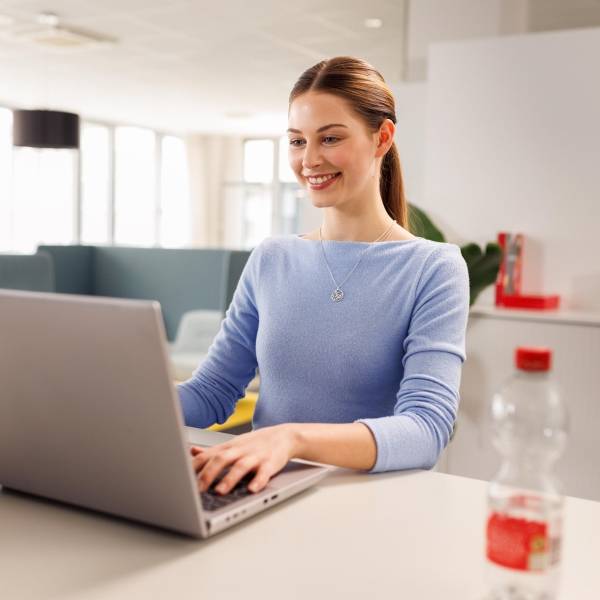 Ein Trainee, eine junge Frau mit fliederfarbenem Oberteil, sitzt im Büro am Laptop und arbeitet. Sie hat einen freudigen Gesichtsausdruck.