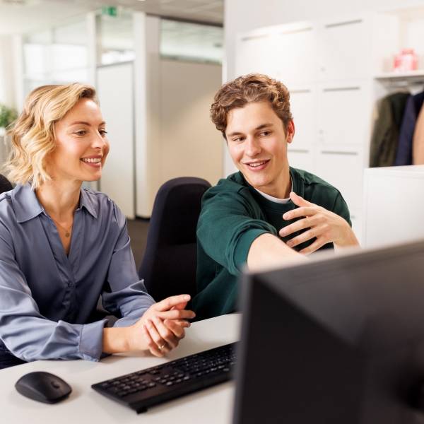 Eine Ausbilderin und ein Student sitzen am Arbeitsplatz vor einem Computer. Die beiden unterhalten sich.