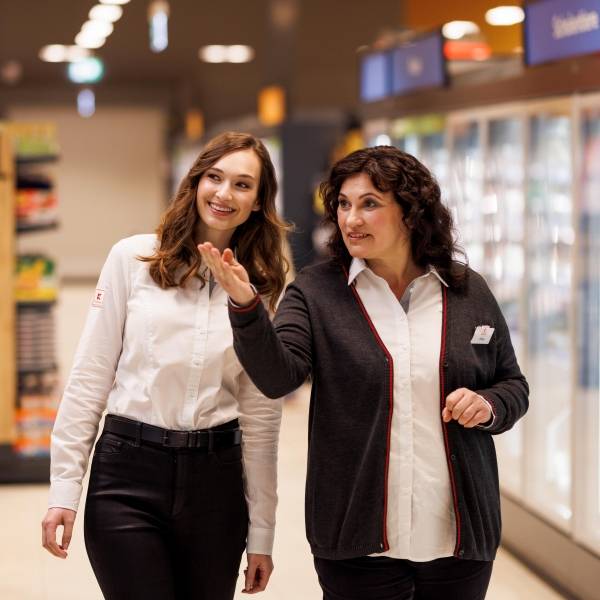 Eine weibliche Führungskraft und eine Mitarbeiterin befinden sich auf einem Rundgang durch eine Kaufland-Filiale. Die beiden sind im Gespräch. Hinter ihnen sind übermannsgroßen Kühlschränken zu sehen.