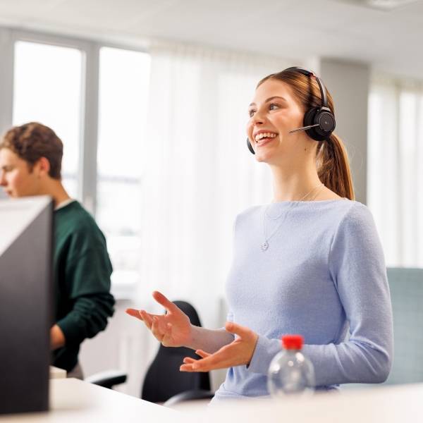 Ein weiblicher Azubi in hellblauem Pullover hat ein Headset auf und führt im Stehen ein Gespräch. Im Hintergrund sieht man halb verdeckt von einem Computer einen weiteren Azubi.