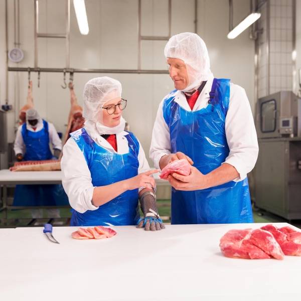 Ein Ausbilder und ein Azubi in blau-weißer Schutzkleidung stehen an einem Tisch in der Azubi-Werkstatt für die Fleischzubereitung. Die Azubi prüft Fleischqualität, während der Ausbilder ihr etwas erklärt.