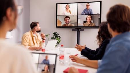 Hybride Zusammenarbeit: 4 Mitarbeiter im Besprechungsraum bei einem Termin mit anderen Kollegen, die über Teams auf einem großen Bildschirm zugeschaltet sind.