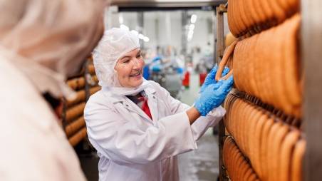 Qualitätssicherung im Kaufland-Fleischwerk: Eine Mitarbeiterin und ein Mitarbeiter in Schutzkleidung vor einem Regalstand mit aufgehangenen Bockwürsten.